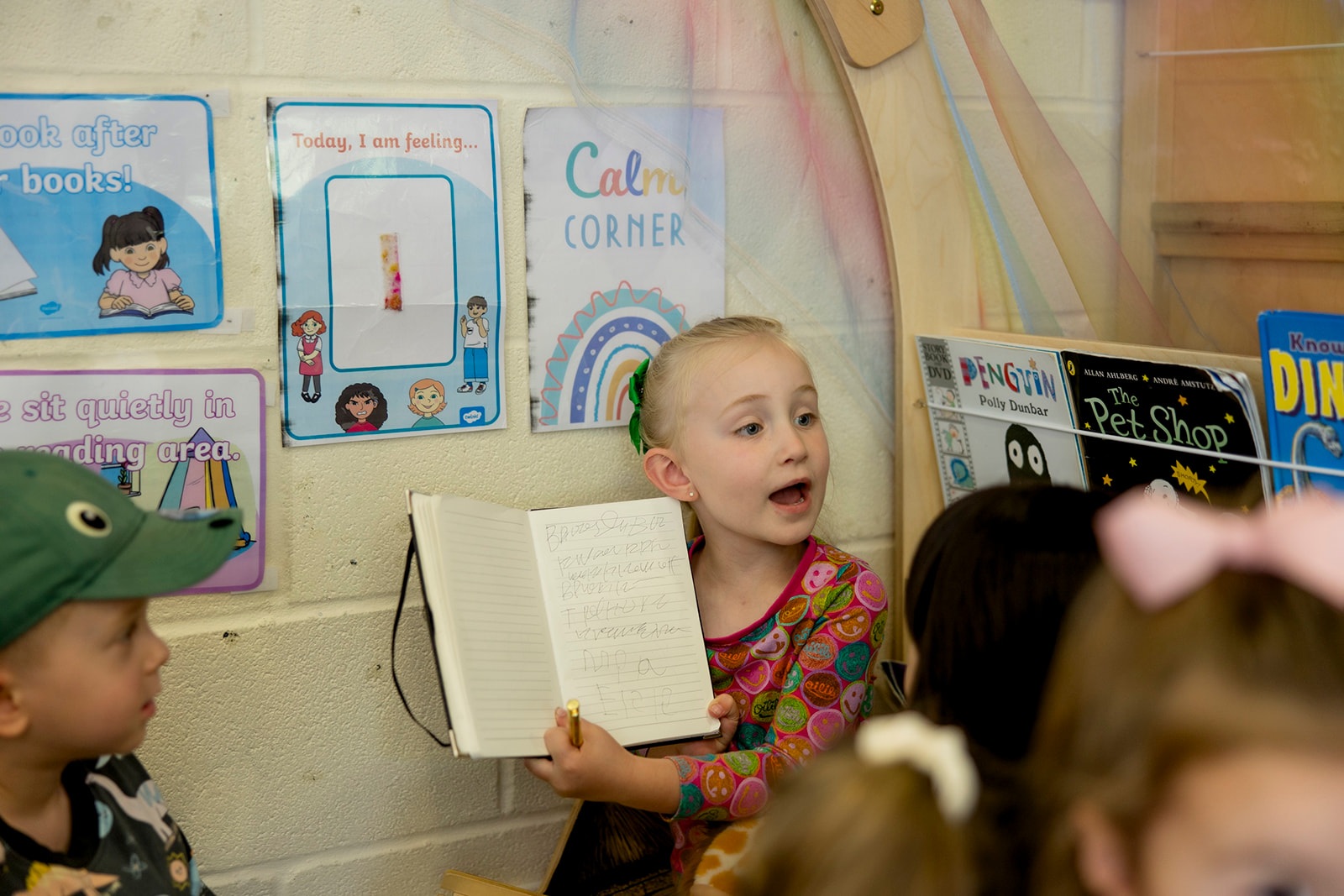 A girl holding up a notebook and reading aloud to other children in the book corner