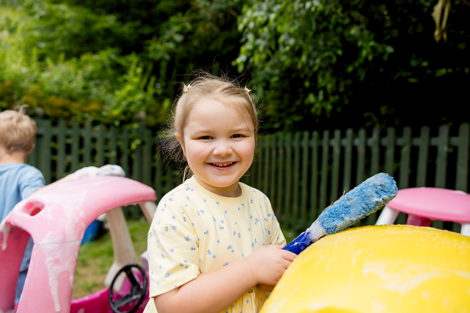A smiling girl brushing soap over a yellow toy car in the garden